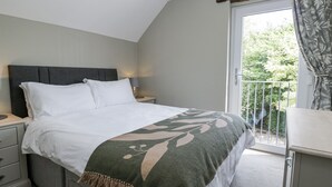 A bedroom with a bed, bedside tables, a nightstand, a window with a view of greenery, and a balcony with a railing.