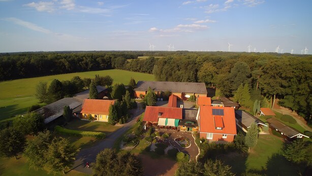 An aerial view of a rural property with multiple buildings, a paved driveway, and wind turbines in the background. - Apartment 'Haarkotten' with Private Terrace, Shared Garden and Wi-Fi (Steinfurt)