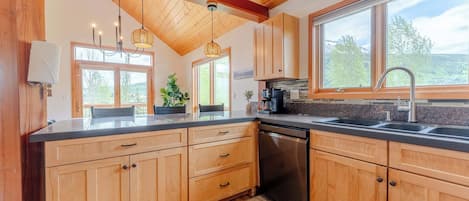 A kitchen with wooden cabinets, a black countertop, a dishwasher, and a window with a view of trees.