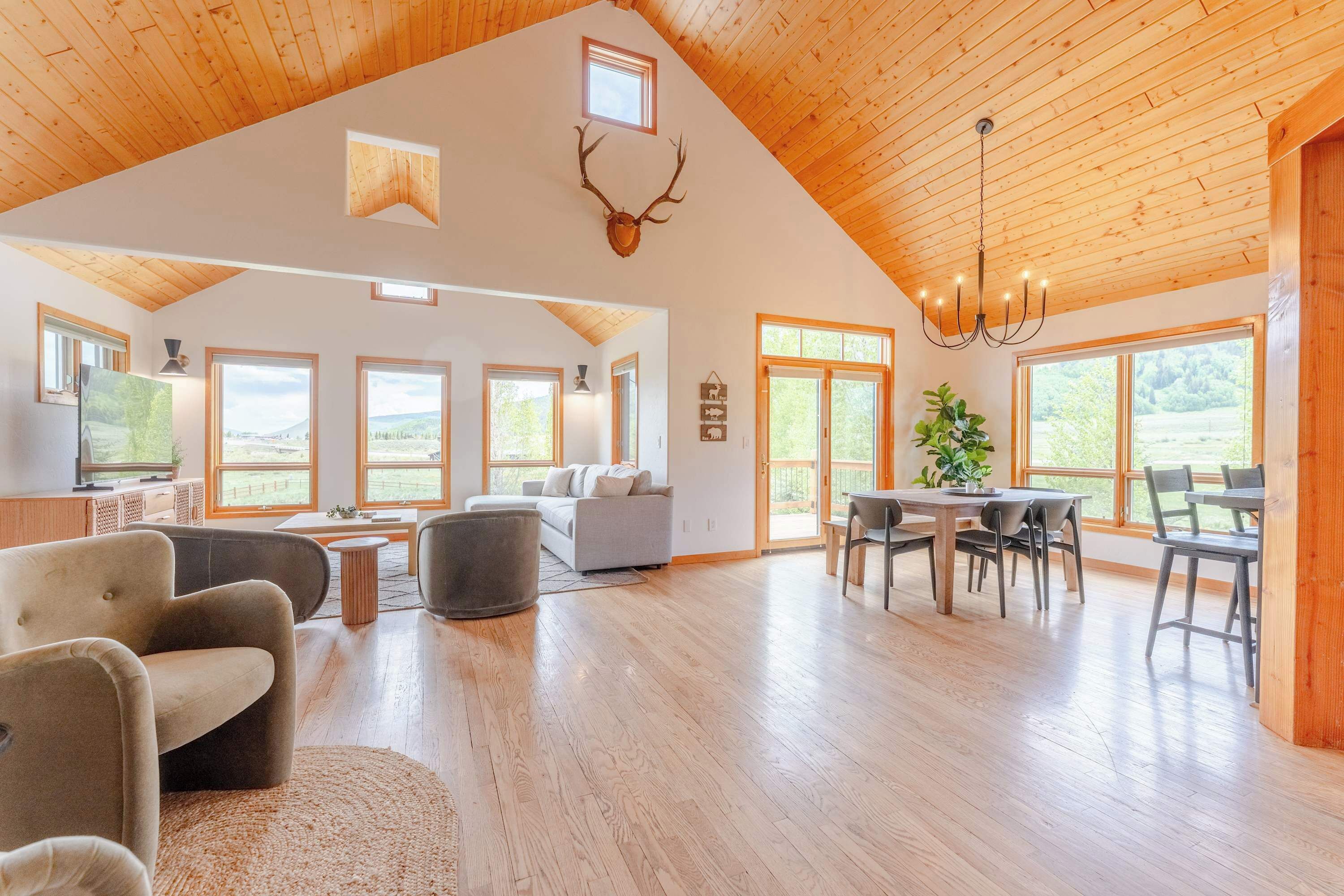 A spacious living area with a high ceiling, wooden beams, a chandelier, a dining table with chairs, and a television on a cabinet.