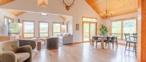A spacious living area with a high ceiling, wooden beams, a chandelier, a dining table with chairs, and a television on a cabinet.