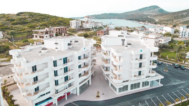 A modern apartment complex with multiple buildings, a parking area, and a view of the sea and hills in the background. - Captain's Residence (Ksamil)