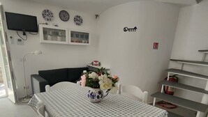 A dining area with a checkered tablecloth, white chairs, a vase of flowers, a television, and a kitchen area with shelves and a fire extinguisher. - Casa Pitturini comfortable apartment in quiet area 500 m from the sea (Marciana Marina)