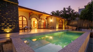A poolside area with a stone wall, a pool, and a covered seating area with a bench and a table. - Lavender Villa by Ebony Stays – Peaceful 3 BHK Getaway in Nashik (Gangavhare)