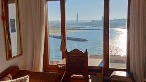A room with a large window overlooking a body of water, a wooden chair, a small table, and a sofa with floral cushions.