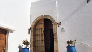A white building with a wooden door and a stone archway leading to a dark interior. There are two potted plants on the steps and a street lamp on the right.