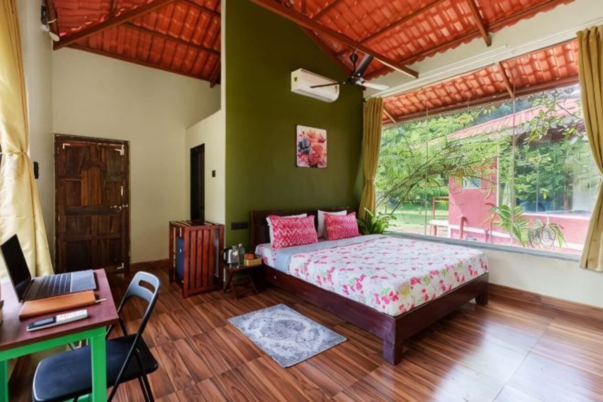 Un dormitorio con una cama, un escritorio con una silla, una ventana con vistas a la vegetación y un ventilador de techo.
