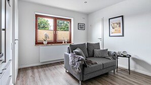 En moderne stue med en grå sofa, et lille sofabord, en radiator, et vindue med rullegardiner og et billede i ramme på væggen.