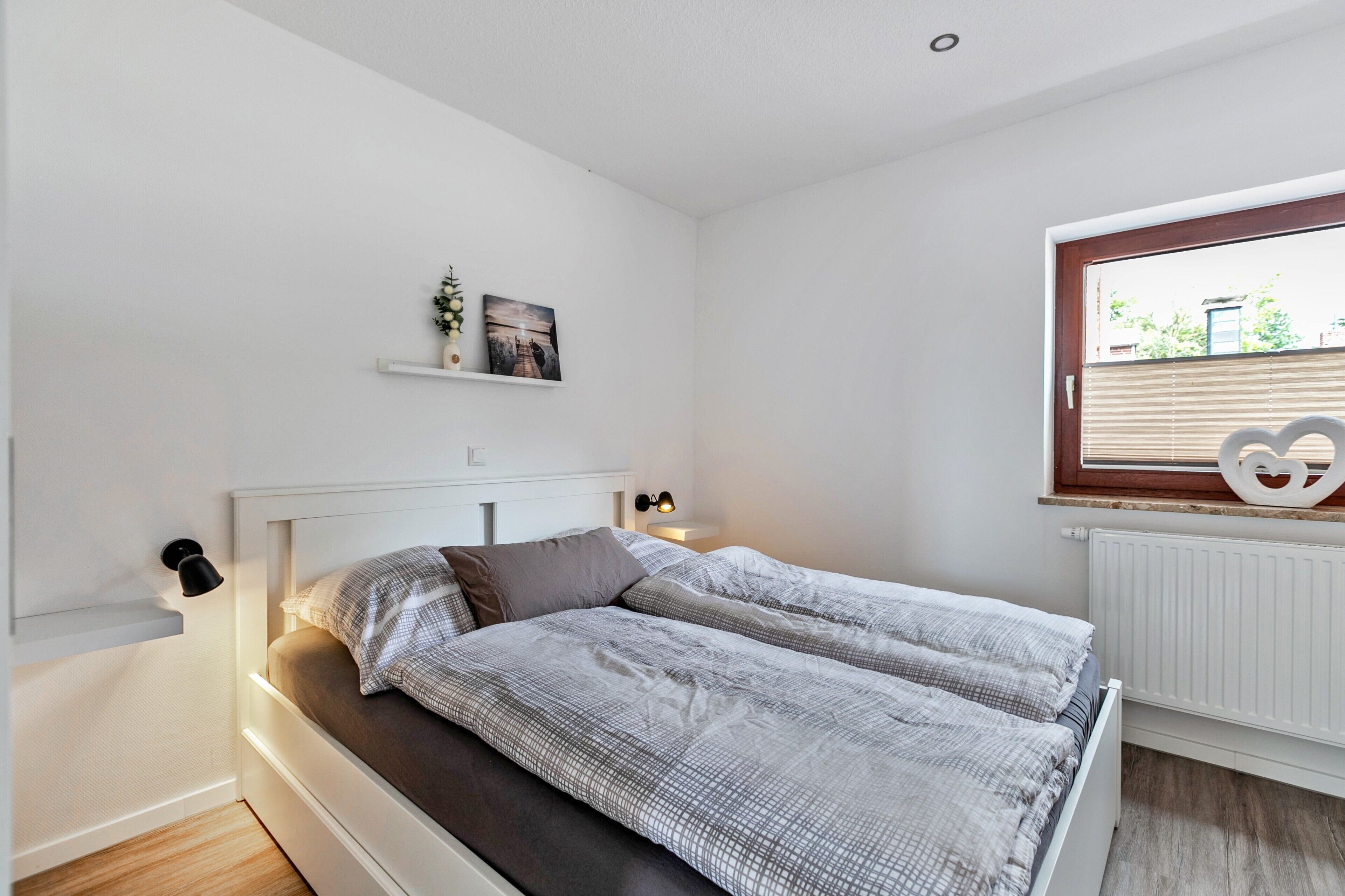 Une chambre à coucher comprenant un lit, des tables de chevet, une fenêtre avec des stores, un radiateur et une étagère ornée d’objets décoratifs.
