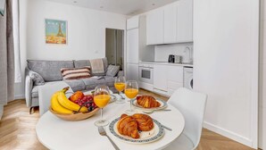 A modern living room with a grey sofa, a dining table with croissants and fruit, and a kitchen in the background. - Charming 1BR Near Eiffel Tower - Sleeps 4 (Paris)