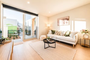 A modern living room with a sofa, coffee table, and a view through large sliding doors to an outdoor area with plants and a fence.