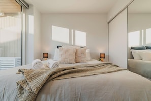 A modern hotel room with a large bed, two bedside lamps, a window with blinds, and a view of a balcony.