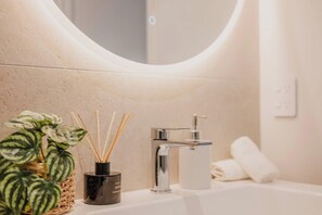 Un baño con un espejo redondo, un grifo moderno, una planta en maceta y un difusor de aroma en un frasco.