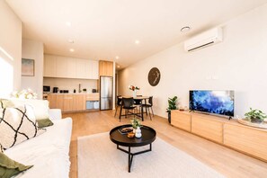 A modern living room with a sofa, coffee table, and a TV. There is a kitchen in the background with cabinets and a refrigerator. - Central 1BR Apartment | Full Kitchen + Patio (Christchurch)