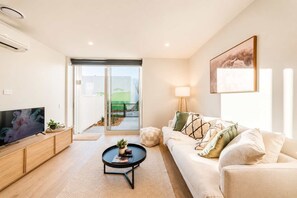 A modern living room with a flat-screen TV, a sofa, a coffee table, and a sliding glass door leading to an outdoor area. - Central 1BR Apartment | Full Kitchen + Patio (Christchurch)