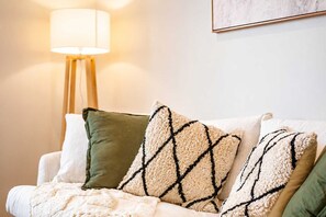 Un sofá con una manta blanca y almohadones estampados, una lámpara de pie y un cuadro en la pared.