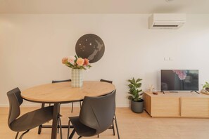 A modern living room with a round wooden dining table, black chairs, a flat-screen TV on a wooden cabinet, a wall-mounted air conditioner, and a decorative wall piece.