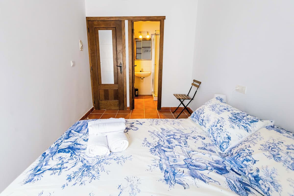 Un dormitorio con una cama, una silla, una puerta de madera y un baño visible a través de una puerta abierta.