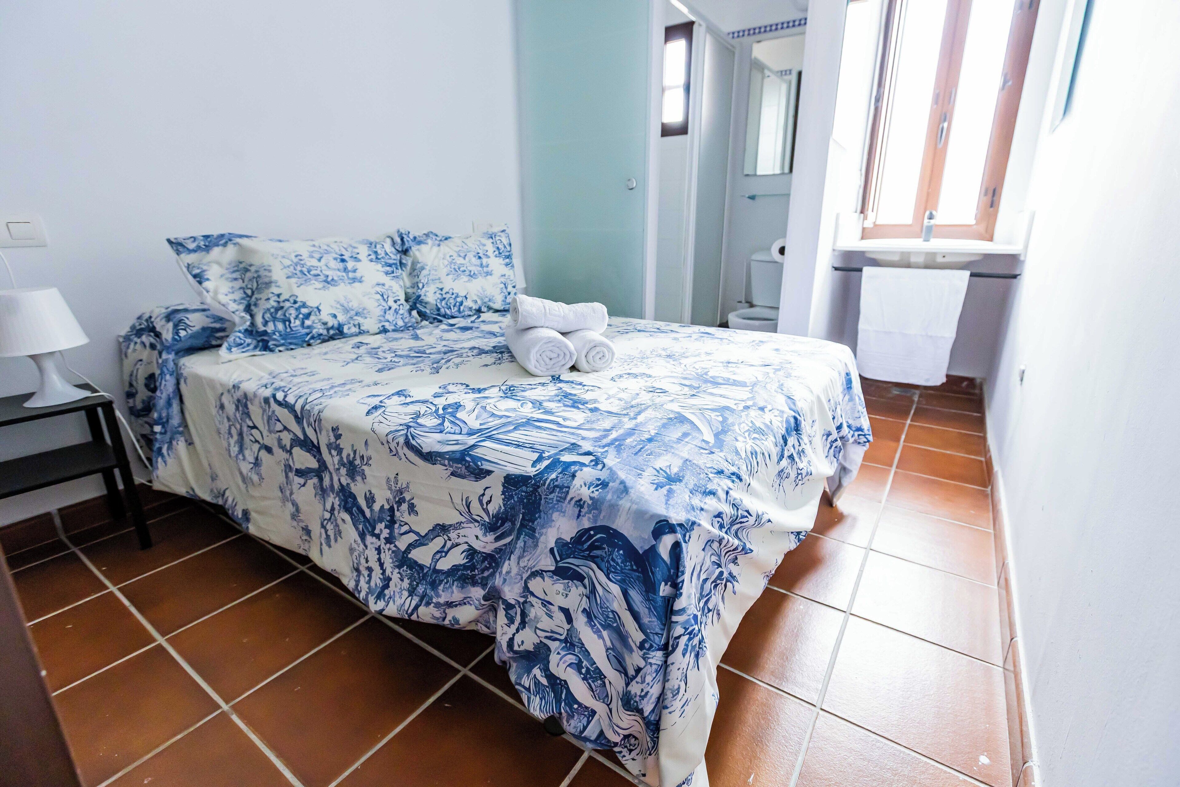 Una cama con una colcha azul y blanca estampada, una mesita de noche con una lámpara y un baño visible al fondo.