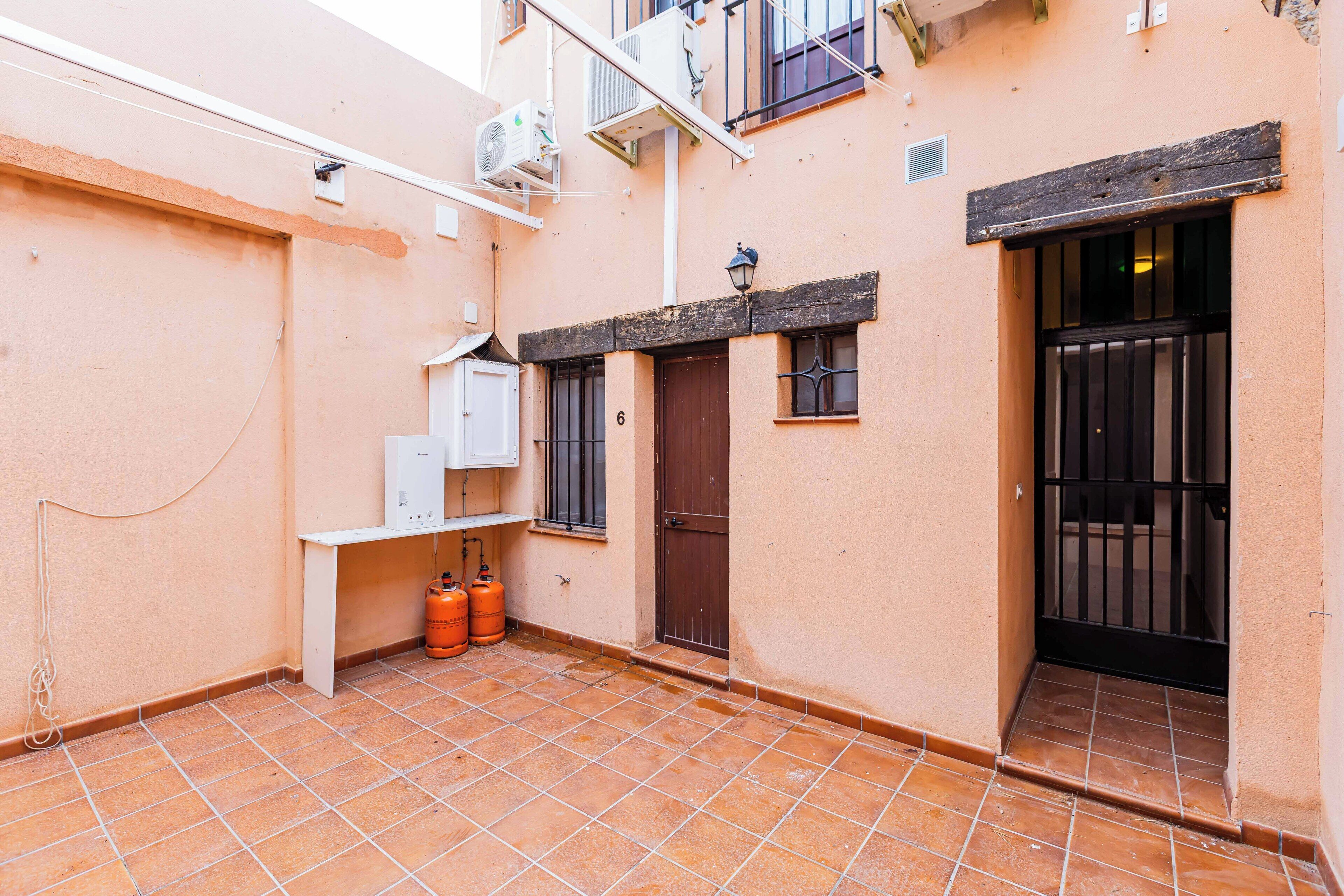 A small courtyard with a tiled floor, a white cabinet, two orange gas cylinders, a brown door, and a black gate.