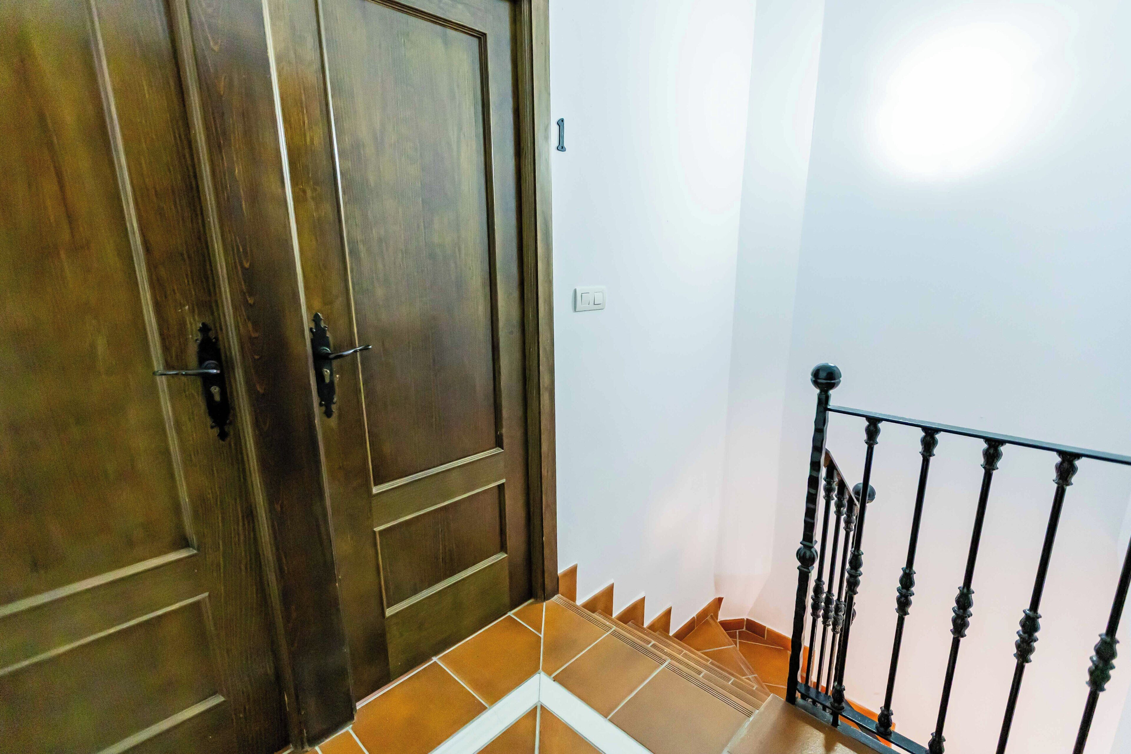 Una puerta de madera con un picaporte metálico, una pared blanca con un interruptor de luz y una barandilla de metal negro.