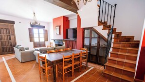 Amplia sala de estar con una mesa de comedor, sillas, un sofá y una televisión. Una escalera con barandilla de madera conduce a un nivel inferior.