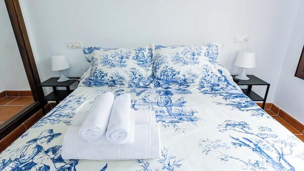 Una cama con ropa de cama estampada en flores azules, dos lámparas de noche y toallas dobladas con cuidado sobre la cama.
