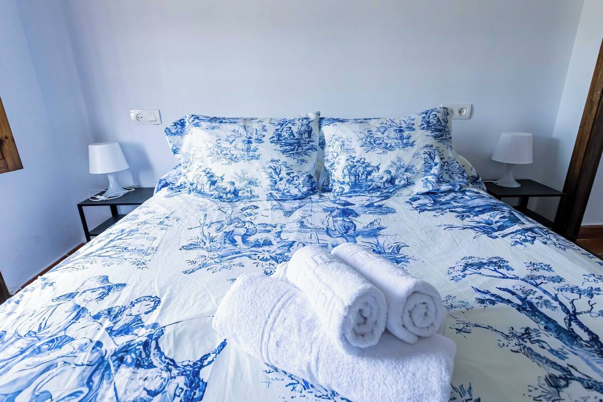 Una cama bien hecha con una colcha azul y blanca estampada con flores, dos almohadas blancas y dos toallas blancas enrolladas sobre la cama.
