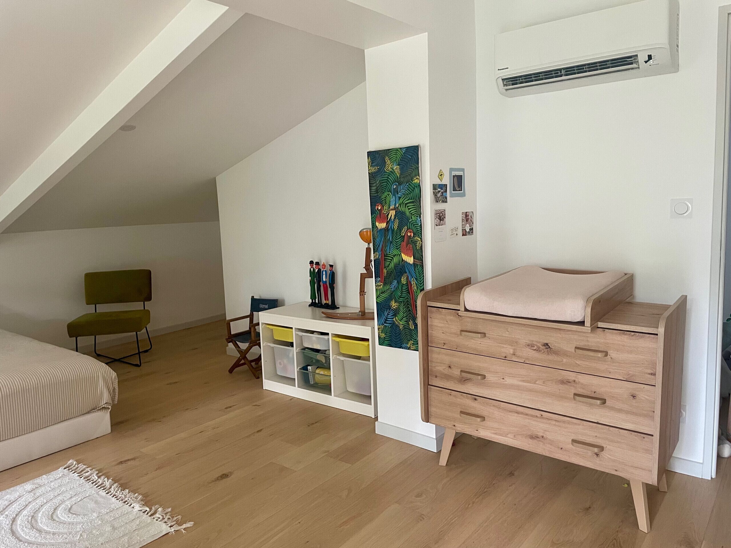 Ein Schlafzimmer mit schrägverdeckter Decke, einem Bett, einem Schminktisch, einem Schreibtisch mit Stauraum, einem Sessel und einem farbenfrohen Kunstwerk an der Wand.