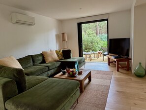 Ein Wohnzimmer mit einem grünen Sofa, einem Couchtisch mit Früchten, einem Fernseher und Blick auf eine Terrasse mit Gartenmöbeln.