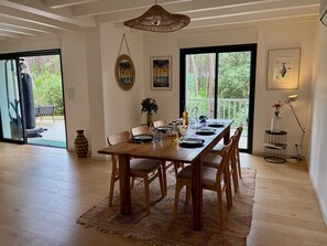 Un coin repas avec une table en bois dressée pour quatre personnes, entourée de chaises, d’un luminaire suspendu et offrant une vue à travers une grande fenêtre.