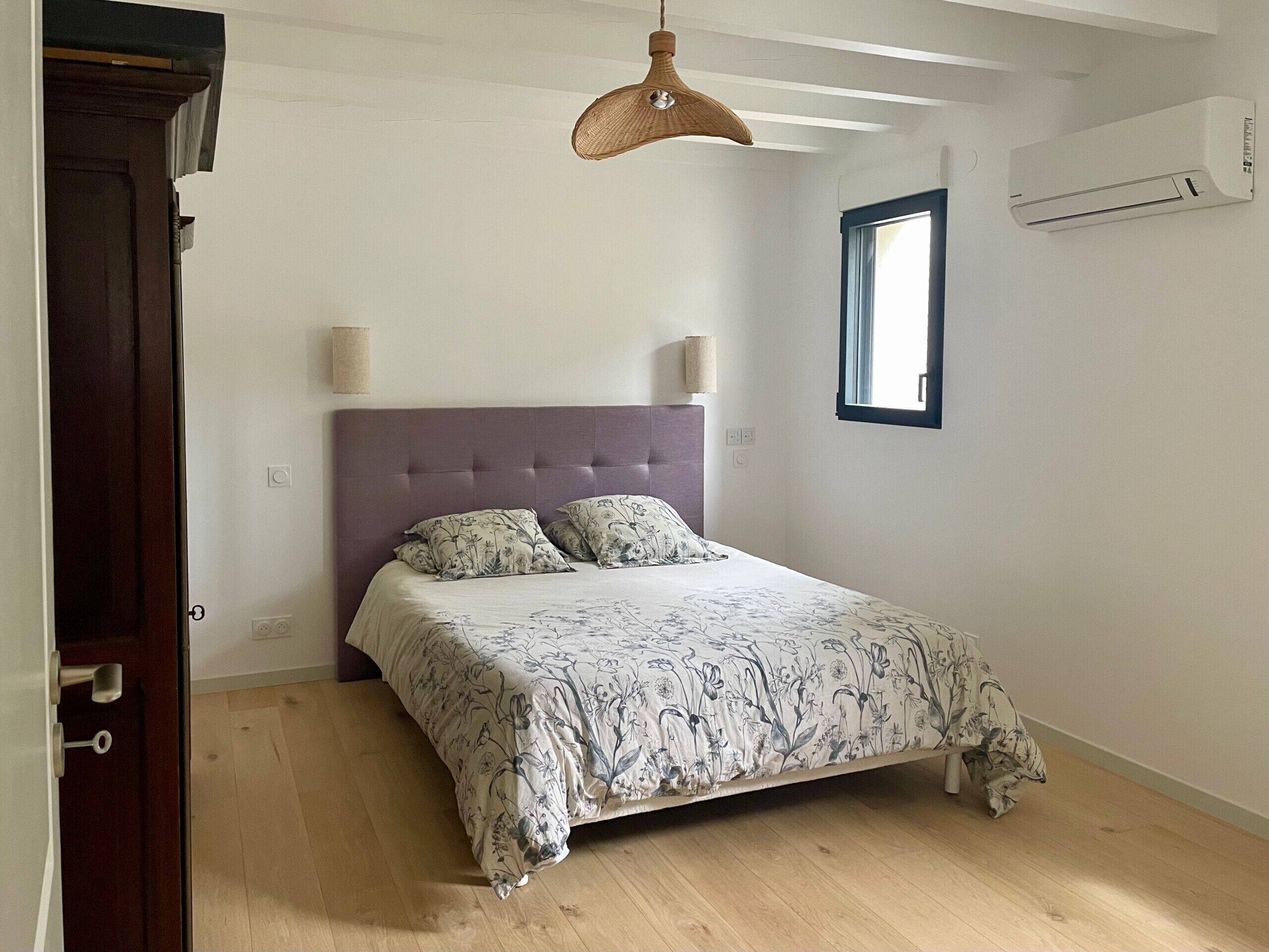 A bedroom with a bed, wooden floor, white walls, a window, and an air conditioning unit.