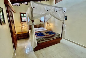 A four-poster bed with a canopy, a wooden headboard, and a patterned bedspread. There are two bedside lamps and a small wooden table beside the bed.