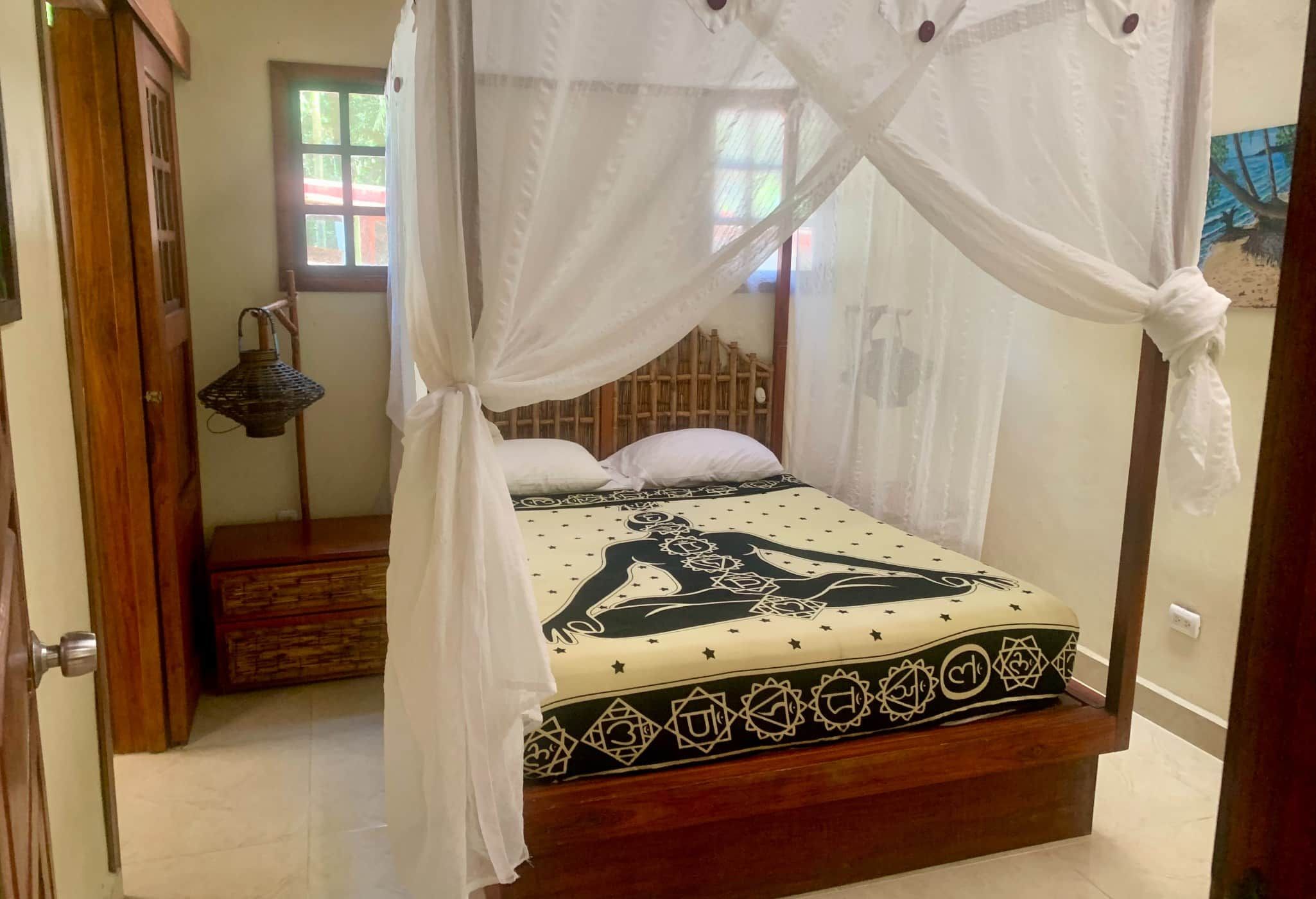 A room with a bed under a canopy, a wooden headboard, a bedside lamp, and a window with curtains.