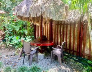 Un espace extérieur aménagé avec une toiture de chaume, comprenant une table et deux chaises, entouré de verdure et délimité par une clôture en bois.