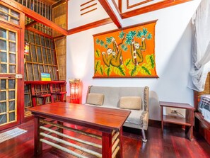 Une pièce avec une table en bois, un canapé, un tableau représentant des singes dans un arbre et une fenêtre avec des stores en bambou.