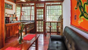 A cozy living area with a wooden bench, a black leather sofa, and a dining table with a red placemat. - Villa #2 - 1 Bedroom with private Garden (Puerto Viejo de Talamanca)