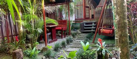 A tropical garden with a wooden bench, a swing, and a thatched roof structure.