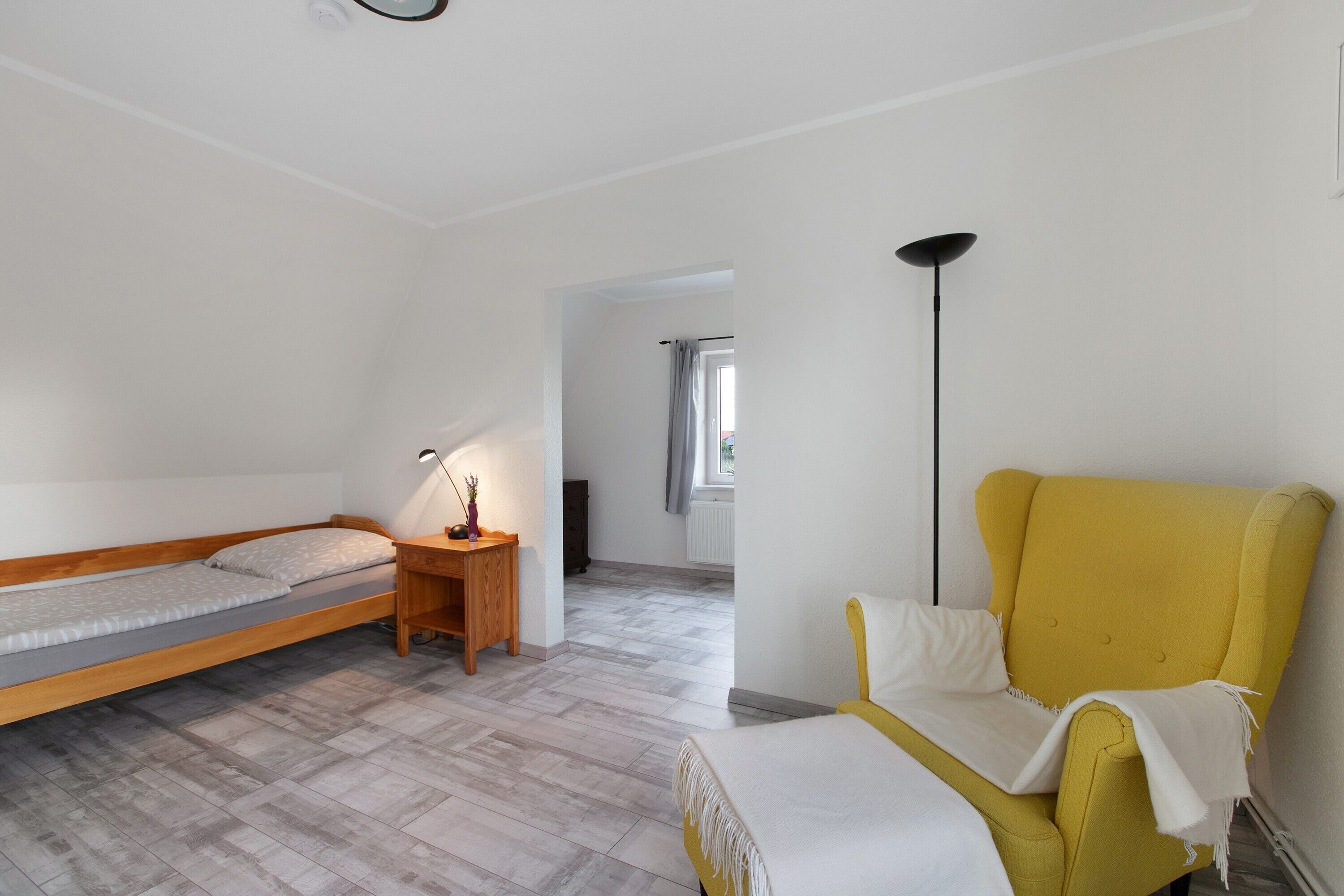 Una habitación con una cama, un sillón amarillo, una lámpara y vistas a través de un puerta abierta.