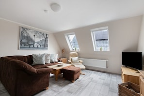 Un salon moderne avec un canapé marron, une table basse en bois, un fauteuil blanc, un téléviseur et une grande fenêtre donnant sur des immeubles.