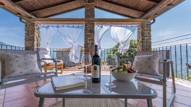A terrace with stone pillars, white curtains, and a table with a bottle of wine, glasses, and a bowl of fruit. - Casa Bandoni, Bagni di Lucca, Lucca and Viareggio (Bagni di Lucca)