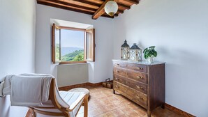 A room with a wooden ceiling, a window with a view of green hills, a wooden dresser with drawers, and a chair with a white cushion. - Casa Bandoni, Bagni di Lucca, Lucca and Viareggio (Bagni di Lucca)