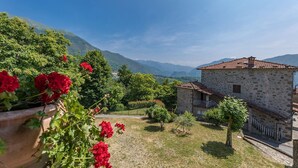 A stone building with a tiled roof, surrounded by a garden with red flowers and trees, set against a backdrop of mountains. - Casa Bandoni, Bagni di Lucca, Lucca and Viareggio (Bagni di Lucca)