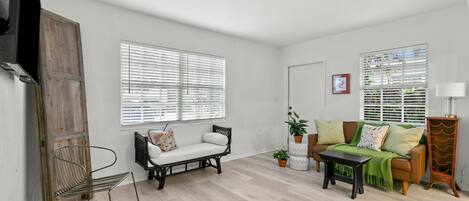Een ruime woonkamer met een bank, een salontafel en een fauteuil. Twee ramen met zonwering, een radiator en een wandgemonteerde televisie.
