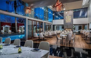 Un vaste espace de restauration intérieur, doté de nombreuses tables et chaises, de grandes fenêtres et offrant une vue sur la piscine.