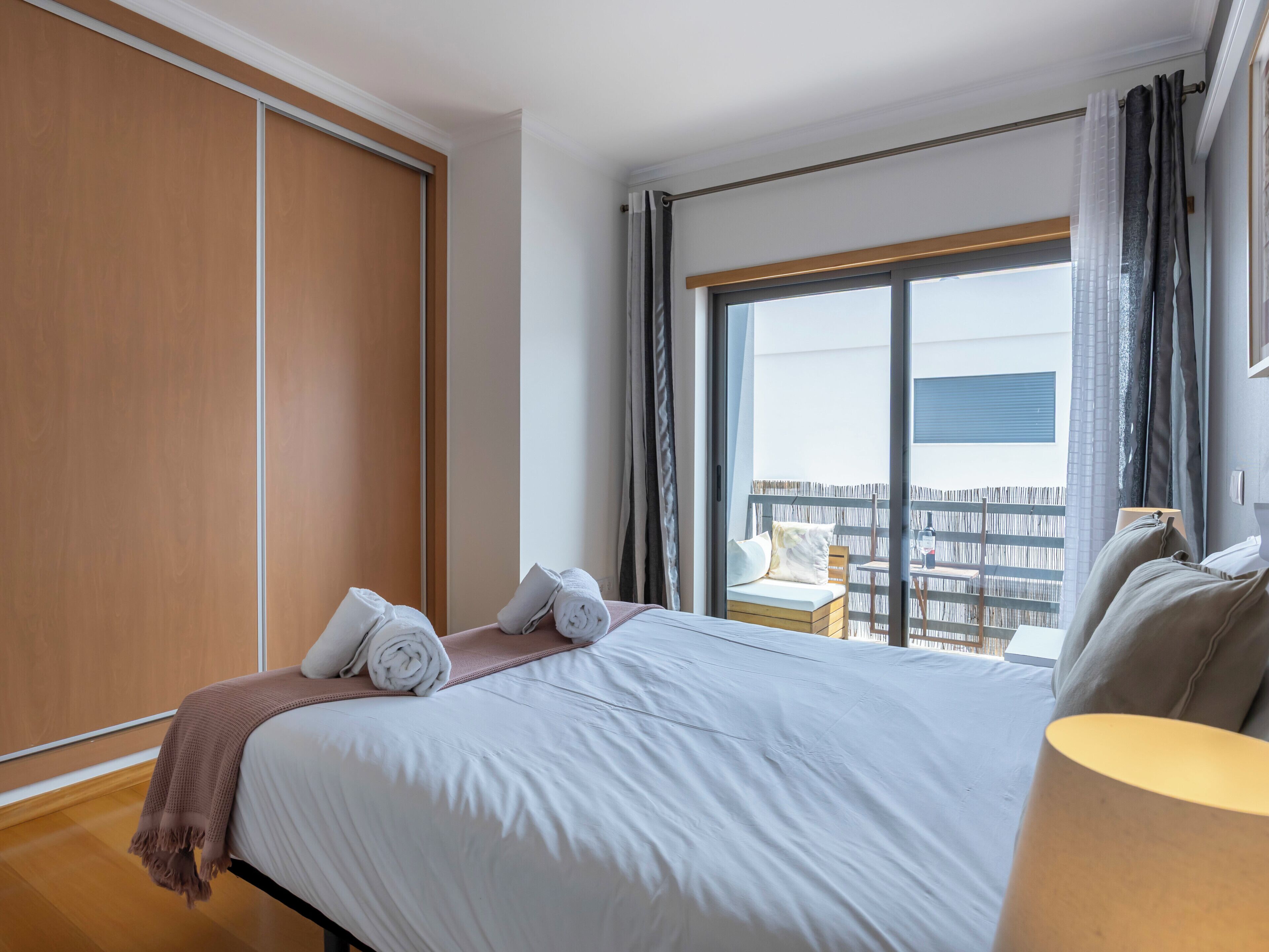 Une chambre d’hôtel avec un grand lit, une armoire en bois, un balcon avec vue et une petite table avec une lampe.