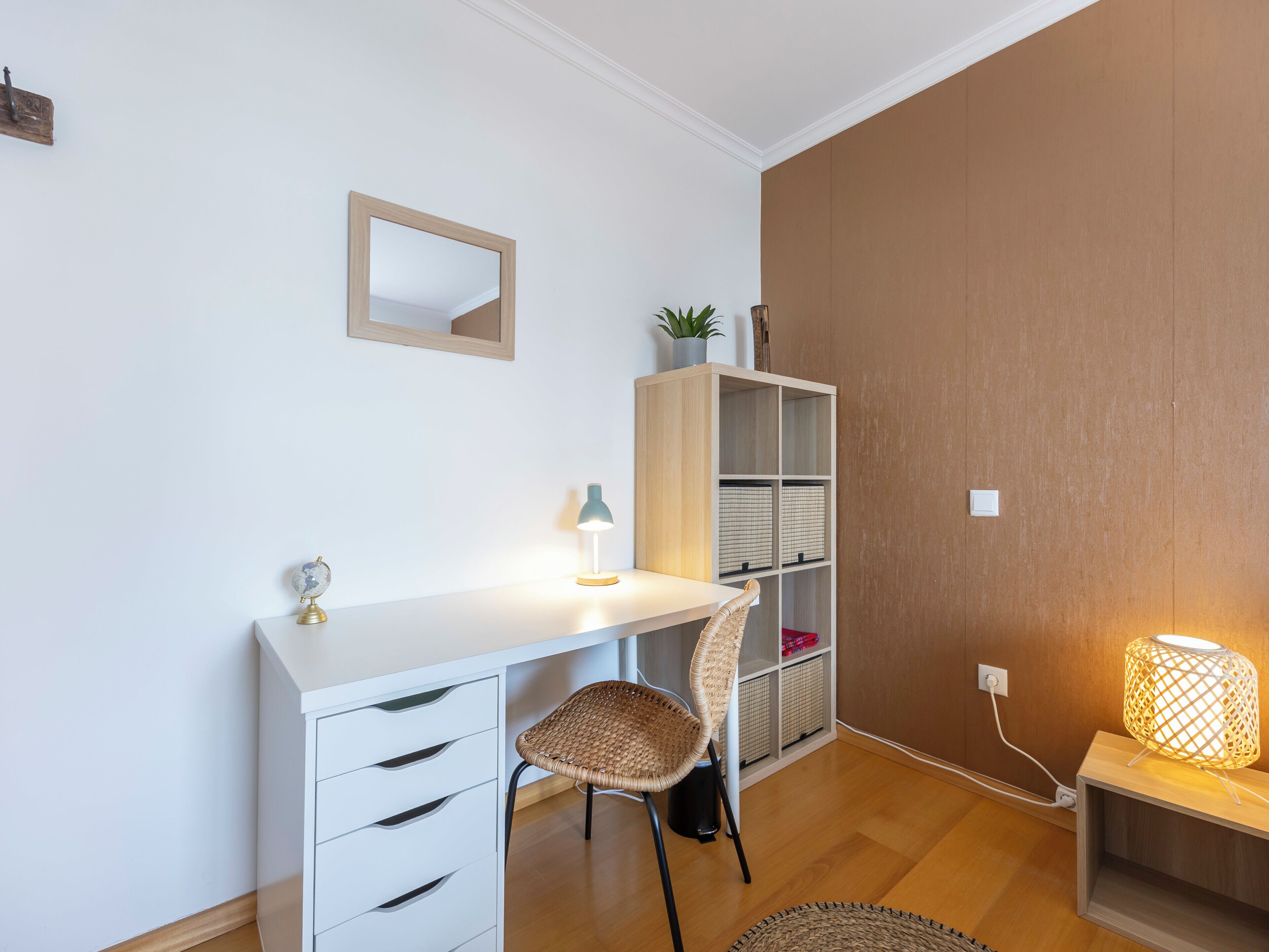 A modern, well-lit room with a white desk, a wicker chair, a bookshelf, and a lamp.