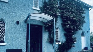 Un bâtiment doté d’une entrée abritée, d’une allée gravillonnée et d’un arbre recouvert de plantes grimpantes.