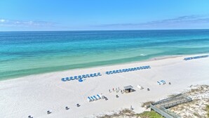 A beach with rows of blue umbrellas and beach chairs, a small building, and a boardwalk leading to the water.