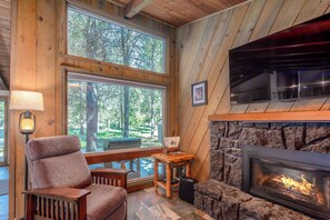 A cozy room with a stone fireplace, wooden walls, a large window with a view of trees, a comfortable chair, and a small table with a lamp. - Sunriver Modern Lodge – Hot Tub & Bikes Included (Sunriver)